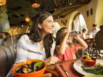 Mum sat next to her daughter who is looking out the windows with binoculars at the animals. They have healthy dishes on the table including a salad. The theming is safari themed.