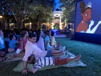 Movies Under the Stars at Disney’s Port Orleans Resort – Riverside Group of guests sat on grass watching a movie at night outside. They're watching The Princess and the Frog