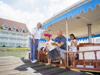 Disney's Grand Floridian Resort & Spa Family getting off of a small boat wearing mickey ears and holding merchandise. In the background there is a white building hotel.