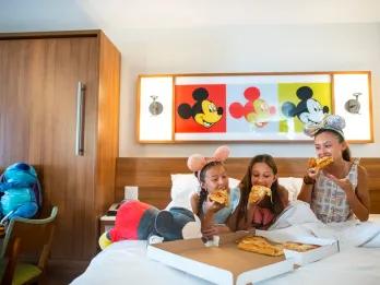 Three young girls sat on a hotel bed eating a large takeaway pizza