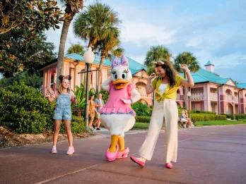 A woman and young girl dancing with Daisy Duck in front Caribbean-style buildings