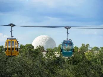 Disney Skyliner at EPCOT Two cable cars, one orange and one blue, moving along cables in front of Spaceship Earth, a giant golf ball looking building.