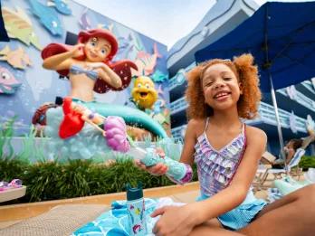 Disney's Art of Animation Resort Girl sat on a sunbed holding an Ariel bubble wand in front of a giant statue of Ariel sat with flounder on the side of the hotel.