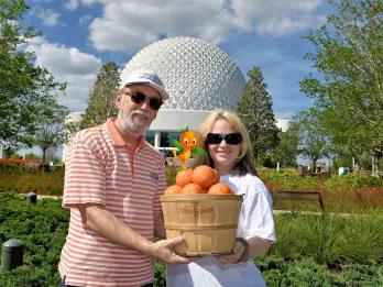 Man and woman holding a basket full of oranges in front of the giant golf ball looking structure and green grass and trees.