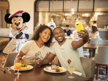 Breakfast à la Art with Mickey & Friends, Topolino's Terrace Flavors of the Riviera at Disney's Riviera Resort Man taking a selfie with his girlfriend smiling at a restaurant. In the background Minnie Mouse is posing with an artist outfit on.