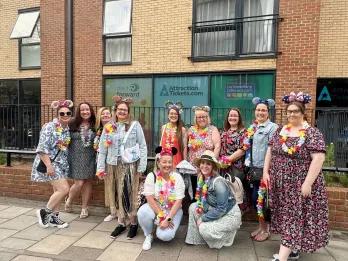 AttractionTickets.com Office Group of women stood outside of an office building with windows with images of Mickey Mouse and the AttractionTickets logo on it. The women are dressed up for the summer party wearing florals and Mickey ears.
