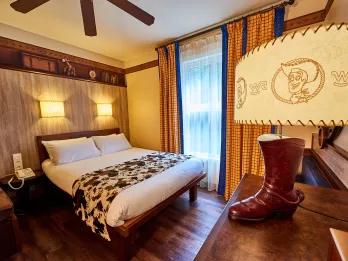 Disney Hotel Cheyenne Standard room with wooden floor, yellow curtains and a double bed. It is themed like the wild west with cow print blankets over the bed and a lamp with woody from toy story on it and a cowboy boot.