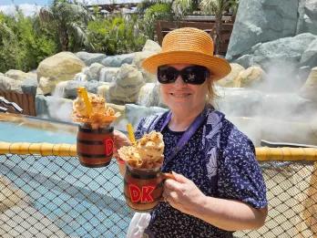 Bubbly Barrel’s DK Crush Float with Mug Woman in theme park holding two barrel themed mugs filled with drinks, whipped cream, sprinkles and more.