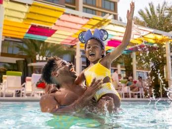 Dad lifting his daughter up in the pool as she laughs and raises her hand. In the background there is a colourful terrace for guests to sit and relax on.
