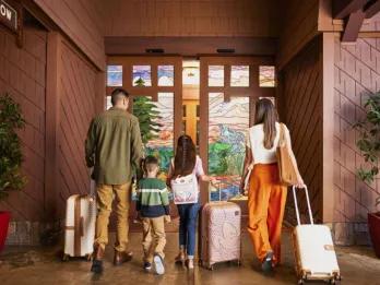Family of 4, parents, a son and a daughter entering sliding doors with amazing art on it, pulling along suitcases.
