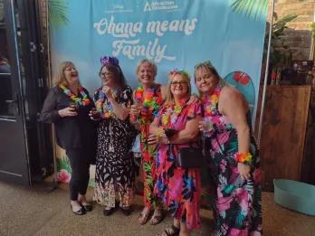 Five women wearing floral dresses a floral hula necklaces holding drinks in front of a backdrop saying "Ohana means family"
