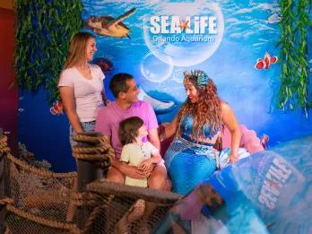 Mermaid with blue tail and seashells in her hair meeting a family including parents and a small kid. In the background there is a SEA LIFE sign.