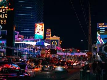 Cars lined up on the road waiting in a traffic jam and there are billboards and flashing lights surrounding them.
