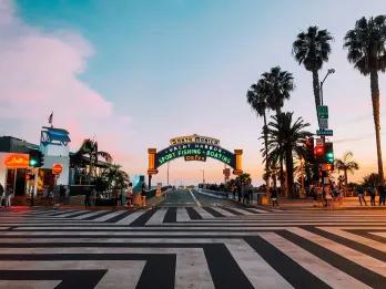 Santa Monica Zig zag black and white lines on the floor leading up to a large sign for Santa Monica pier lighting up as the sun sets. There are palm trees next to it.