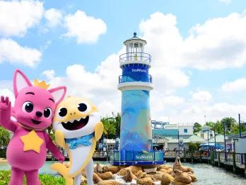 Two mascot animals, one pink dog creature wearing a yellow crown. They are stood in front of a blue lighthouse on the rocks.