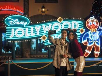 A couple taking a selfie in front of a neon sign for Disney Jollywood Nights