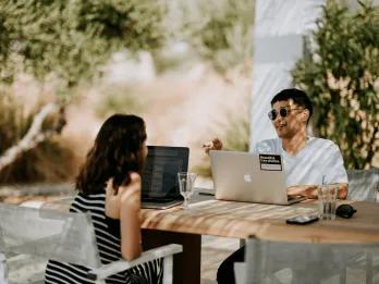 A woman and man sat at a wooden table outside talking in the sun with their work laptops open.
