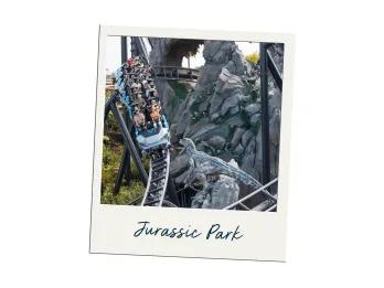 Jurassic Park People riding an intense roller coaster which loops around grey rocks making sharp turns at high speeds. There's a dinosaur roaring at the people passing by.