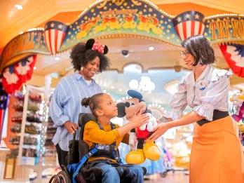 A Disney cast member handing a Mickey Mouse plush to a child in a wheelchair. Her mum is standing behind her