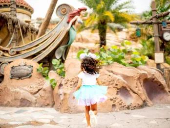 A young girl in a tutu and Minnie ears standing in front of a shipwreck with Ariel on the front