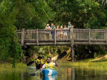 People on a wooden bridge watching people kayak along a river