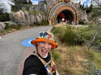 Sarah - Jump the Queue Podcast Host Woman in a bucket hat which has text on it saying Monsters and cartoon art on it. She's stood with her mouth open in excitement in front of one of the Epic Universe portals.