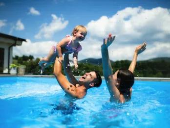 A family enjoying a high-energy "darecation" moment in a sunny outdoor pool, with water splashing as a father lifts a young child