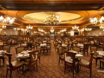 Elegant dining room interior of Silver Spur Steakhouse at Disneyland Park, featuring cowhide chairs, ornate chandeliers, rich wood panelling and a grand domed ceiling with a Western-inspired décor