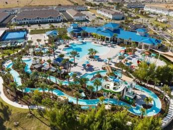 An aerial drone view of a resort water park featuring a large swimming pool, lazy river, water slides, sports courts, and a main clubhouse building with a blue roof.