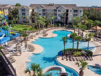 Aerial overview of a lagoon-style resort swimming pool, water slides, and multi-story vacation condo buildings under a clear sky.