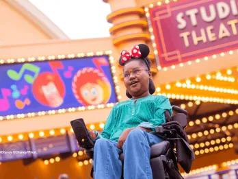 Guest in a wheelchair wearing Minnie Mouse ears smiling beneath the Studio Theater marquee at Production Courtyard, Disney Adventure World