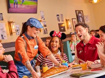 Server presenting food to a laughing group of guests at the Sports Bar & Lounge presented by Coca-Cola, Disney Village