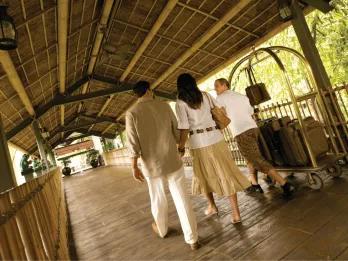 A couple walking along a covered bamboo walkway at Loews Royal Pacific Resort while a hotel staff member pushes a luggage cart