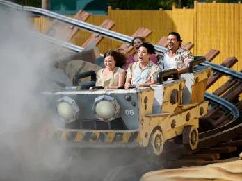 Riders smiling and cheering aboard a rustic mine cart roller coaster emerging from a cloud of mist