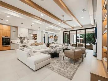 A spacious modern open-concept living room and kitchen featuring wooden ceiling beams, a large white sectional sofa, and floor-to-ceiling glass patio doors