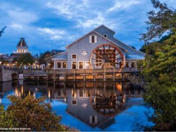 A large wooden water wheel attached to a building reflected in a river at twilight
