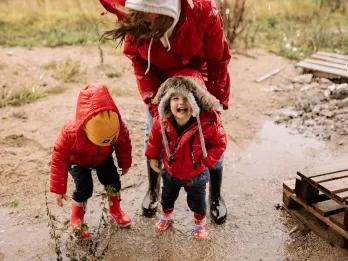 Two young children in red raincoats laugh and play in a muddy puddle while an adult in a red coat bends down beside them, illustrating a rainy day out with the family