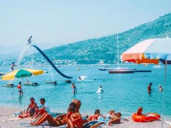 Sunbathers and swimmers enjoying a busy Mediterranean beach with a water slide and parasols.