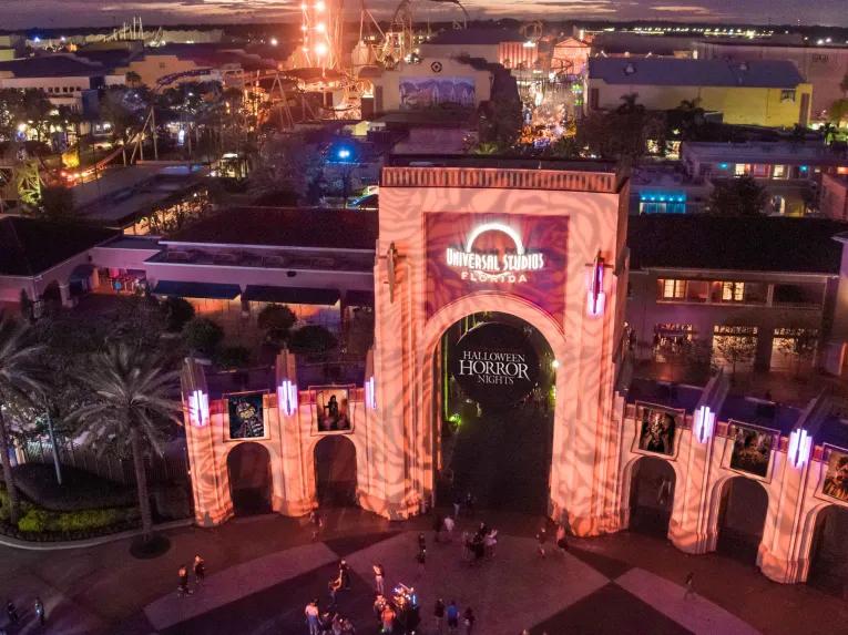Bird’s eye nighttime view of Universal Studios Florida arches with a circular Halloween Horror Nights banner in the middle and the park illuminated behind.