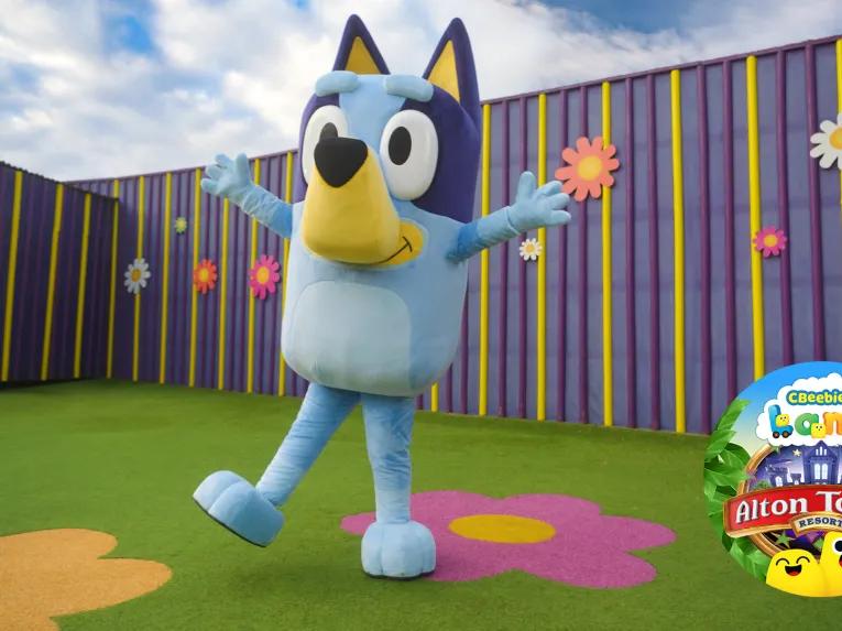 Big blue dog called Bluey with arms extended open and the Alton Towers CBeebies Land logo in the bottom right corner