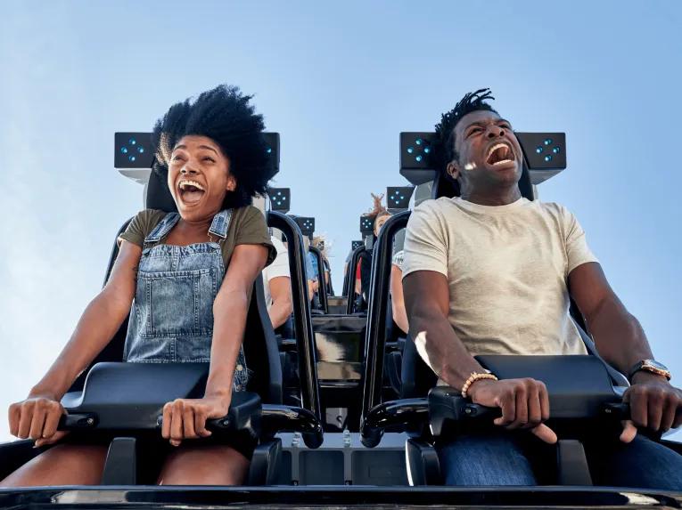Two guests on black rollercoaster clutching onto the lap bars holding them into the car. Both are smiling whilst screaming.