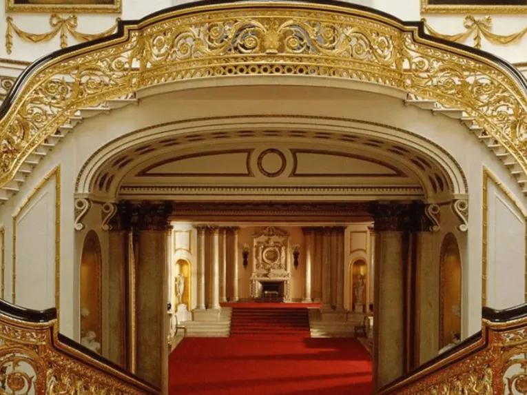 Golden and wooden bannisters lining two staircases which wrap around and join to create on large staircase. There is bright red carpet and white walls lined with gold details. There is a large open hallway with a fireplace at the end.