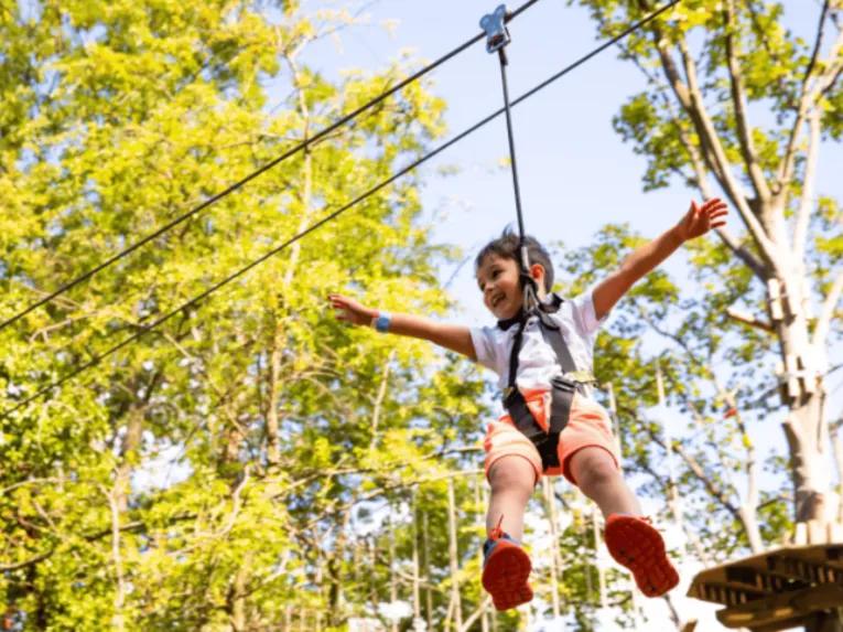 Little boy on zip line dangling in the tree tops held by harness. He has a smile on his face and his arms reaching out wide.