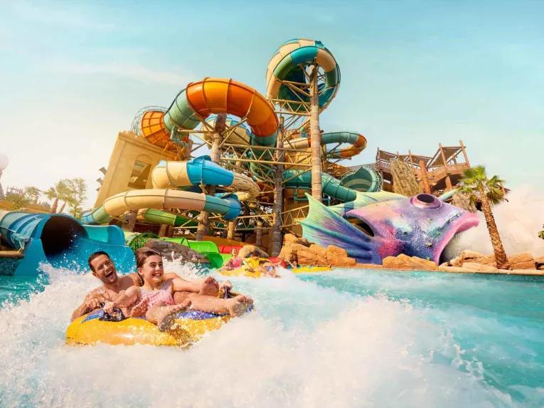 A couple enjoys a thrilling water slide ride at Yas Waterworld, Abu Dhabi, with colorful slides and vibrant scenery in the background.